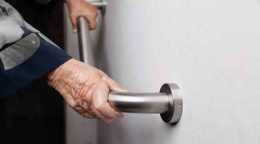 An elderly homeowner holds a steel handrail with both hands as they walk down the steps inside of their home.