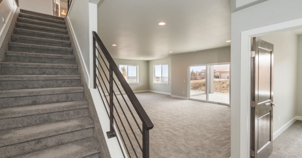An empty downstairs area in a house with gray carpeting, white trim, a sleek metal handrail, and sliding glass doors.